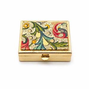 Può includere: Piccola scatola per pillole rettangolare color oro con coperchio incernierato. Il coperchio è decorato con un colorato motivo floreale nei toni del verde, blu, rosso e giallo. La scatola ha un design semplice ed elegante.