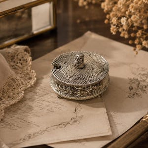 Puede incluir: Un detallado recipiente de filigrana plateada con tapa, sobre papel envejecido con diseños florales. El recipiente tiene una pequeña cuchara y está rodeado de objetos vintage, lo que sugiere un tema de decoración antigua.