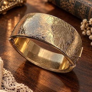 Può includere: Un ampio bracciale rigido incernierato in metallo color oro caratterizzato da un intricato motivo floreale in rilievo su tutta la superficie esterna. Il bracciale presenta una finitura lucida e un meccanismo di chiusura sicuro sul lato.