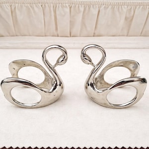 May include: Two silver swan-shaped napkin rings. The napkin rings are displayed inside a brown box with a cream-colored interior. The swans have a smooth, reflective surface and a circular opening for the napkin. The box has a gold clasp.