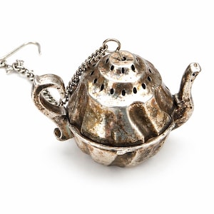 Op de afbeelding: Zilverkleurige thee-infuser in de vorm van een kleine theepot. Het heeft een scharnierende basis, een tuit en een handvat. Een ketting en haak zijn bevestigd voor eenvoudig gebruik. De bovenkant is geperforeerd met kleine gaatjes.
