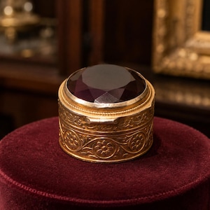 Peut inclure: Une boîte ronde en métal doré ornée d'un motif floral en relief sur les côtés et dotée d'un couvercle à charnière. Le dessus du couvercle est incrusté d'une grande pierre précieuse facettée violet foncé qui reflète la lumière.