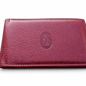 May include: A burgundy leather wallet with a textured finish. The wallet has a stitched edge and a central oval emblem. The emblem features a stylized double "C" logo. The wallet is rectangular and appears closed.