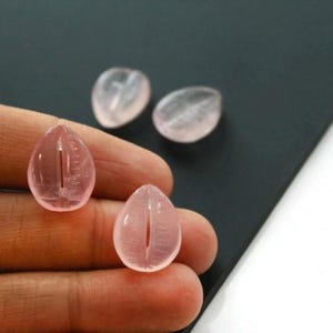 May include: Four translucent, teardrop-shaped rose quartz beads. Two beads are in focus, held by a hand, with a vertical groove. The other two beads are out of focus in the background. The beads are a soft pink color.