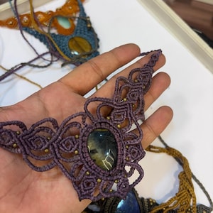 May include: A purple macrame necklace featuring an oval-shaped tiger eye gemstone at the center, surrounded by intricate knotted cord patterns and small metallic beads. The design includes long, thin cords extending from the sides for fastening.