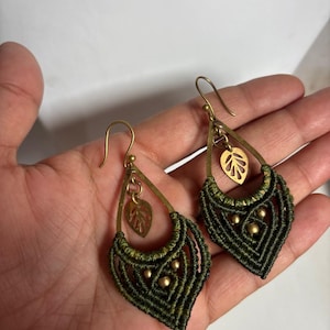 May include: A pair of teardrop-shaped earrings featuring dark olive green macrame cord woven into a geometric pattern. Each earring includes a brass-toned metal frame, a small dangling leaf-shaped charm, and three tiny brass beads embedded in the cord.