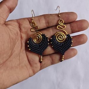 Puede incluir: Un par de pendientes colgantes con una espiral de metal dorado en la parte superior, conectada a una base de macramé negro en forma de abanico. El diseño de cordón negro está decorado con pequeñas cuentas doradas en los bordes y una cuenta en la punta.
