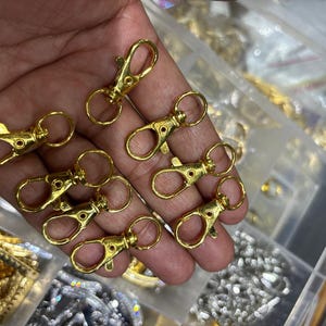 Puede incluir: Un conjunto de cierres de langosta de metal en tono dorado, cada uno con un mecanismo de resorte y un anillo giratorio circular en la base, diseñados para sujetarse a joyas, llaveros u otros proyectos de manualidades para una fijación segura.