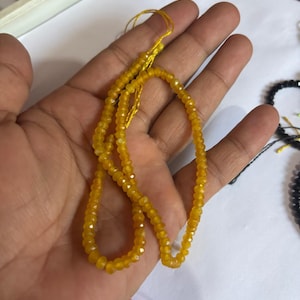 May include: A strand of small, faceted, translucent yellow beads strung on a thin yellow thread. The beads have a reflective, polished surface and are arranged in a loose, circular loop, showcasing their uniform size and vibrant golden-yellow colour.