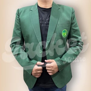 Op de afbeelding: Een effen groene blazer met een enkele knoopsluiting en een golfbaan-embleem op de borst. De blazer wordt gedragen over een zwart t-shirt. De blazer is een klassieke stijl.