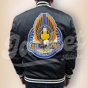 Könnte beinhalten: Schwarze Satin-Bomberjacke mit weiß-schwarzem Streifenbesatz. Auf der Rückseite befindet sich ein großer gestickter Aufnäher mit einem Adler, den Worten "FALL GUY" und "STUNTMAN ASSOC" in einem blau-orangen Design.