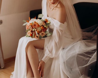 May include: A white floor-length gown with long sleeves, an off-the-shoulder neckline, and a high leg slit. The ensemble includes a sheer veil with scattered pearls, a pearl necklace, and white pointed-toe ankle-strap heels with a bouquet of orange and white flowers.