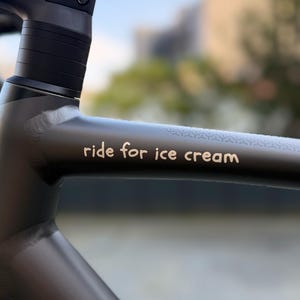 Könnte beinhalten: Nahaufnahme eines dunkelgrauen Fahrradrahmens mit dem Schriftzug "ride for ice cream" in weißer Schrift. Der Rahmen hat ein strukturiertes Design in der Nähe des Oberrohrs. Der Hintergrund ist unscharf.