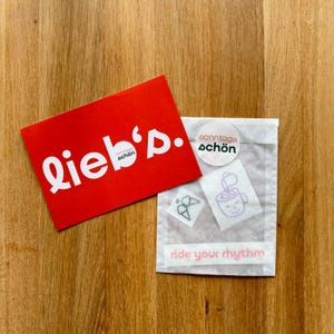 May include: A red card with the word "lieb's" in white, and a small white circle with the words "sonntags sch&ouml;n". Next to it is a small bag with stickers and the words "ride your rhythm".