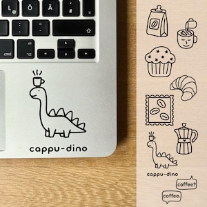 Könnte beinhalten: Ein Laptop mit einem schwarz-weißen Aufkleber eines Dinosauriers mit einer Kaffeetasse auf dem Kopf und dem Wort "cappu-dino" darunter. Zusätzliche Illustrationen zum Thema Kaffee befinden sich auf einer Holzoberfläche.