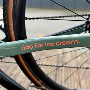 May include: Close-up of a bicycle frame in a muted green color. The frame features the text "ride for ice cream" in a coral color. The image also shows a portion of the wheel and chain.