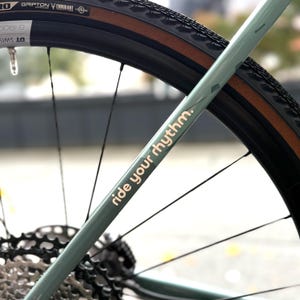 May include: Close-up of a bicycle wheel and frame. The frame is a muted green color with the words "ride your rhythm" in a gold font. The tire is black with a brown sidewall. The image focuses on the rear wheel and gears.