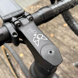 Könnte beinhalten: Nahaufnahme eines schwarzen Fahrradvorbaus mit weißer Eiswaffel-Grafik. Der Vorbau ist am Lenker und einem Fahrradcomputer befestigt. Der Vorbau hat die Zahl "60" und ein Herzsymbol.