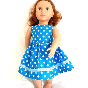 May include: A doll wearing a blue dress with white polka dots. The dress has a sleeveless design and a flared skirt. The doll has red hair and is standing on a white surface.