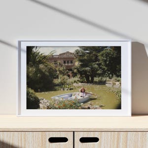 Pode incluir: Fotografia emoldurada de uma pessoa num pequeno barco num lago. O barco é azul claro e a pessoa está a usar um chapéu. A vegetação exuberante rodeia o lago e um grande edifício é visível no fundo.