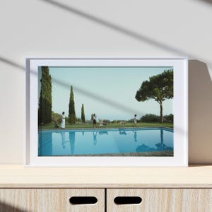 May include: A framed print featuring a serene outdoor scene with a swimming pool. The pool reflects the sky and figures of people and dogs. Tall trees and a lush lawn surround the pool, creating a tranquil atmosphere. The print is displayed on a wooden surface.
