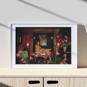 May include: A framed print featuring a woman in a red and white patterned dress, seated on a table laden with fruit. The room has dark wood paneling, red accents, and floral arrangements. A portrait hangs on the wall.
