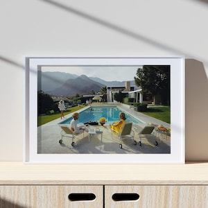 May include: A framed print depicting a mid-century modern scene. Two women relax by a pool, one in a yellow dress, the other in a patterned outfit. The scene includes a pool, lounge chairs, and a mountain backdrop.