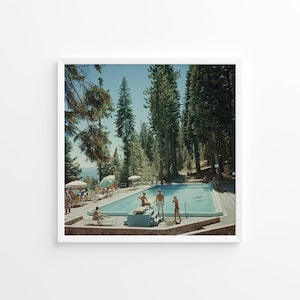 May include: A framed print of a vintage outdoor swimming pool scene. The rectangular pool is surrounded by people, trees, and umbrellas. The water is a light blue color. The print is in a white frame.