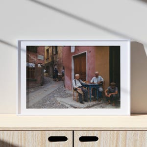 May include: A framed print depicting a European street scene. Three elderly men sit at a small blue table. The buildings are painted in shades of pink, yellow, and brown. A cobbled street leads into the distance.