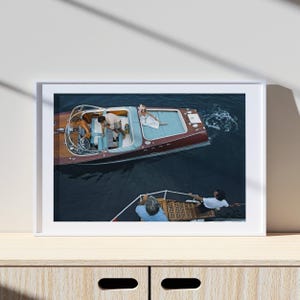 May include: A framed print featuring a vintage-style motorboat with a red hull and wooden deck. Passengers are aboard, and another boat is nearby. The water is dark blue, and the scene evokes a sense of leisure and travel.