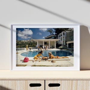 May include: A framed print of a vintage photograph depicting a poolside scene. Two women in swimwear are playing a game near a pool. A group of people are gathered in the background near a large white house. A pink bag and a straw hat are visible.