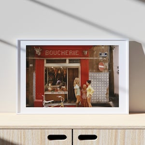 May include: Framed print of a French butcher shop, "Boucherie," with a red facade and a bicycle parked outside. Three people stand in front of the shop. The image is set in a sunny outdoor scene.