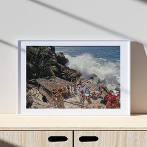 May include: A framed photograph depicts a coastal scene with a crashing wave. People are gathered on a rocky outcrop with a thatched structure adorned with flowers. Red flowers and palm trees frame the scene, with the ocean in the background.