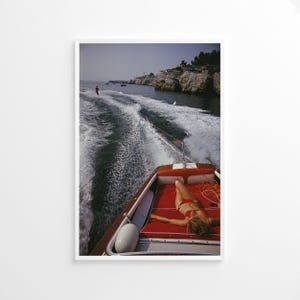 Puede incluir: Fotografía enmarcada de un día soleado en el agua. Una persona en bikini rojo se relaja en un barco acolchado rojo, mientras que un esquiador acuático es remolcado detrás del barco. El barco deja una estela espumosa.