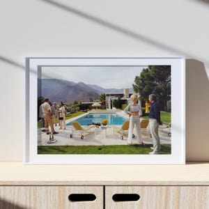 May include: A framed print showing a retro pool party scene. People are gathered near a swimming pool, with sun loungers and a table with flowers. The background features mountains and a mid-century modern house.