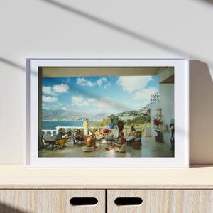 May include: A framed print depicting a group of people socializing on a patio overlooking a scenic coastal view. The scene includes a table with flowers, chairs, and individuals in various attire, set against a backdrop of mountains and a blue sky.