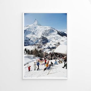 May include: A framed print showing a snowy mountain scene with people enjoying winter activities. The Matterhorn mountain is in the background. People are skiing and relaxing in the snow. The sky is a clear blue.