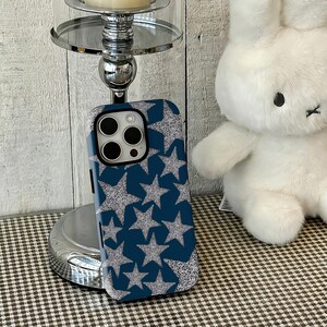 Op de afbeelding: Een blauwe telefoonhoes met een zilveren glitter sterrenpatroon. De hoes staat naast een witte knuffelkonijn en een zilveren kandelaar. De achtergrond is een licht houten paneel.