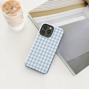 May include: A light blue and white gingham patterned phone case. The case is on a white surface next to a glass of iced coffee and an open book.