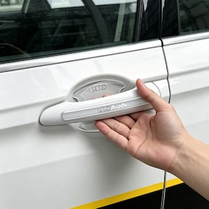 May include: A white car door handle cover featuring the Audi logo and text. The accessory is designed to fit over the existing handle, with a chrome-accented insert behind the handle grip that also displays the Audi brand emblem.