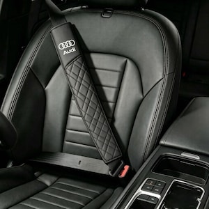 May include: A black seat belt cover featuring a quilted diamond pattern and the white Audi logo. The cover is attached to a black seat belt, which is positioned diagonally across a black leather car seat with horizontal stitching details.