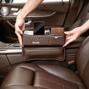 Pode incluir: Um organizador de frestas para assento de carro em couro marrom com o logotipo da Audi em prata. O organizador possui vários compartimentos contendo um smartphone, uma carteira pequena e um chaveiro, projetado para caber entre o console.