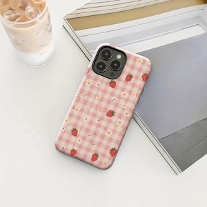 Op de afbeelding: Een telefoonhoesje met een roze ruitpatroon, met rode aardbeien en kleine witte bloemen. De hoes heeft een zwarte cameramodule en ligt op een wit oppervlak.