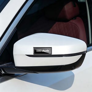 Puede incluir: Un espejo lateral de vehículo blanco con un emblema rectangular negro que muestra el logotipo y el texto de Audi en blanco. La carcasa del espejo tiene una sección superior blanca brillante y un borde inferior negro.