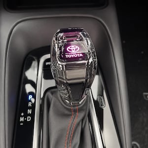 May include: A clear, faceted crystal gear shift knob featuring the Toyota logo and brand name illuminated in purple. The knob is attached to a black leather boot with red stitching, positioned above a gear selection panel with P, R, N, and D settings.