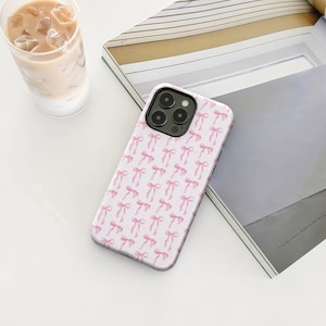 Op de afbeelding: Een witte telefoonhoes met een herhaald patroon van roze strikken. De hoes ligt op een wit oppervlak naast een glas ijskoffie en een gedeeltelijk zichtbaar open boek.
