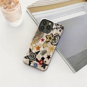 May include: A decorative phone case with a celestial theme. The case features a beige background with gold, black, and red star designs, a small disco ball, and a planet graphic. The case is next to a magazine and a glass of iced coffee.