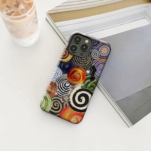 Peut inclure: Une coque de téléphone colorée avec un motif de spirales de différentes couleurs, notamment orange, vert, noir et blanc. La coque est posée sur une surface blanche, à côté d'un verre de café glacé et d'un livre.