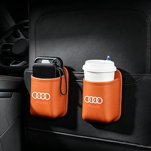 Könnte beinhalten: Zwei orangefarbene Auto-Sitzorganizer aus Leder mit weißem Audi-Logo. Der linke Organizer enthält ein schwarzes Smartphone, während der rechte einen weißen Einweg-Kaffeebecher mit Deckel hält. Diese Aufbewahrungstaschen sind am Sitz befestigt.