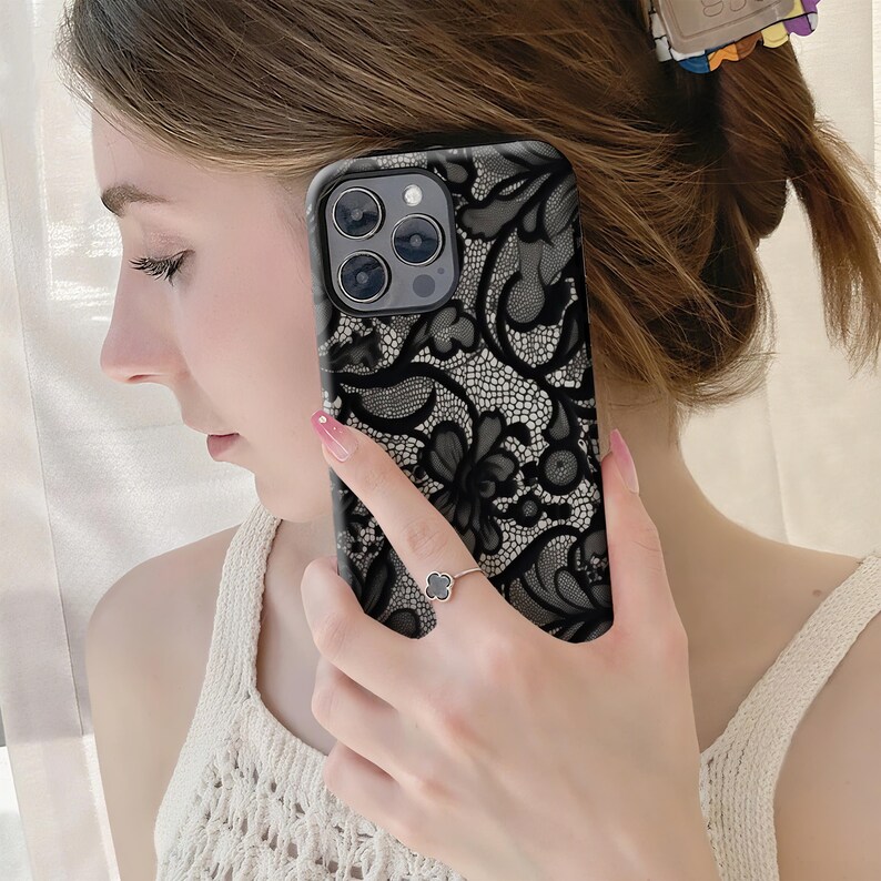 May include: Black lace phone case with a floral design. The case is held by a person. The case has a black and white floral lace pattern. The phone case is a protective accessory for a smartphone.